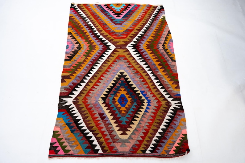 K&ouml;nnte beinhalten: Ein farbenfroher, geometrischer Teppich mit einem Rautenmuster. Der Teppich weist eine Vielzahl von Farben auf, darunter Rot, Blau, Gelb und Braun. Das Design umfasst Zickzack- und Dreiecksformen. Der Teppich ist rechteckig.