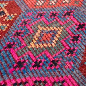 May include: A woven textile featuring a dense geometric pattern with a prominent central diamond motif. The fabric displays a vibrant color palette of deep red, bright magenta, turquoise, light blue, and yellow threads in a textured weave.