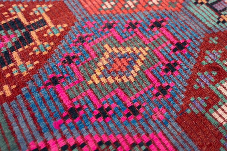 May include: A woven textile featuring a dense geometric pattern with a prominent central diamond motif. The fabric displays a vibrant color palette of deep red, bright magenta, turquoise, light blue, and yellow threads in a textured weave.