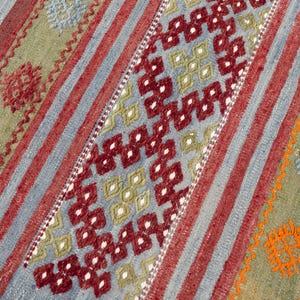May include: A colorful, patterned rug with a geometric design. The rug features a central diamond pattern in red and white, framed by stripes of red, blue, and green. The rug's edges have a pink fringe.