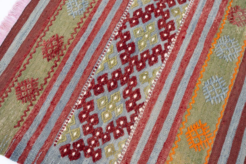 May include: A colorful, patterned rug with a geometric design. The rug features a central diamond pattern in red and white, framed by stripes of red, blue, and green. The rug's edges have a pink fringe.