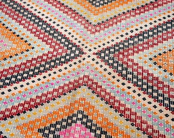 May include: Close-up of a colorful woven textile with a geometric pattern. The design features diagonal stripes in shades of orange, red, black, and blue, with small square accents. The background is a light beige color.