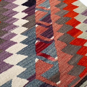 Puede incluir: Primer plano de una colorida alfombra tejida con un patr&oacute;n geom&eacute;trico. La alfombra presenta un dise&ntilde;o repetitivo de diamantes y tri&aacute;ngulos en tonos de rojo, naranja, morado, gris y blanco. La alfombra tiene un borde con flecos.
