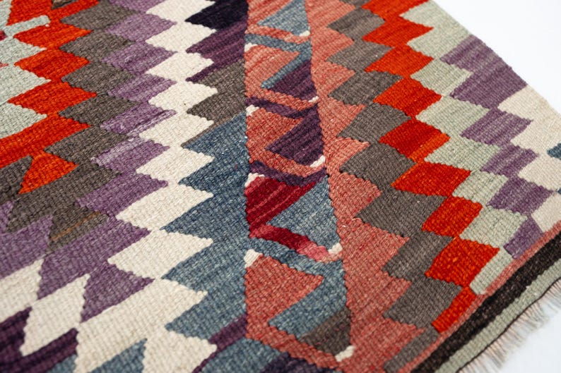 Puede incluir: Primer plano de una colorida alfombra tejida con un patr&oacute;n geom&eacute;trico. La alfombra presenta un dise&ntilde;o repetitivo de diamantes y tri&aacute;ngulos en tonos de rojo, naranja, morado, gris y blanco. La alfombra tiene un borde con flecos.