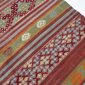 May include: A woven rug with a geometric pattern. The rug features diagonal stripes in shades of red, gray, and green. Intricate diamond and square patterns are woven throughout the design, with accents of orange and white.