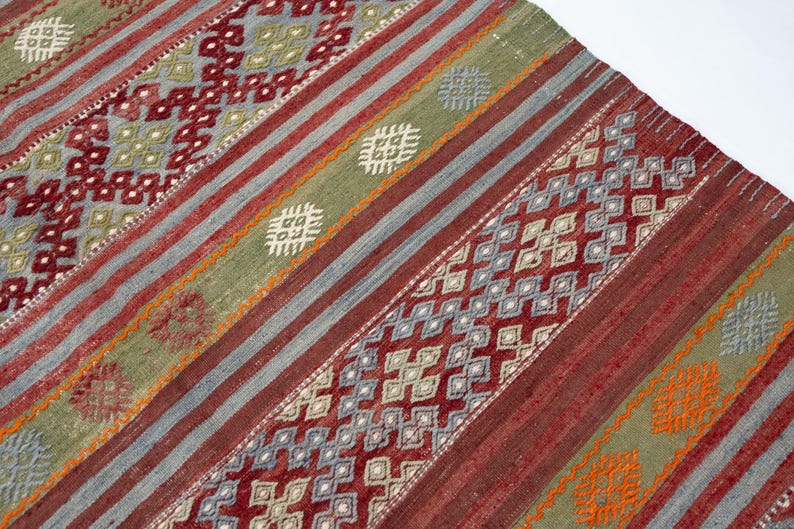 May include: A woven rug with a geometric pattern. The rug features diagonal stripes in shades of red, gray, and green. Intricate diamond and square patterns are woven throughout the design, with accents of orange and white.