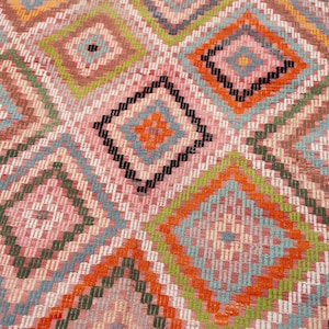 May include: Close-up of a colorful, geometric patterned rug. The rug features a repeating diamond pattern in shades of pink, orange, green, blue, and black. The design is intricate and detailed, with a woven texture.