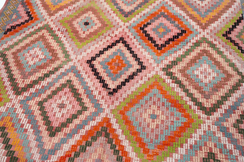May include: Close-up of a colorful, geometric patterned rug. The rug features a repeating diamond pattern in shades of pink, orange, green, blue, and black. The design is intricate and detailed, with a woven texture.