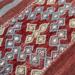 May include: Close-up of a woven textile with a geometric pattern. The design features repeating diamond shapes in shades of red, blue, and white. The background is a deep red, and the overall texture appears soft and slightly worn.