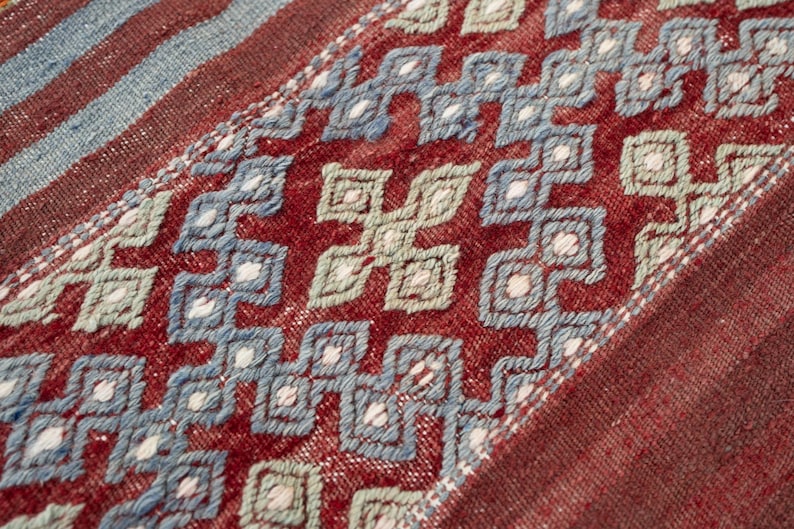 May include: Close-up of a woven textile with a geometric pattern. The design features repeating diamond shapes in shades of red, blue, and white. The background is a deep red, and the overall texture appears soft and slightly worn.