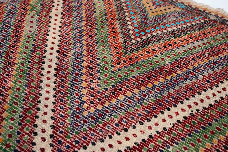 May include: Close-up of a colourful, patterned rug with a geometric design. The rug features a mix of red, green, blue, and yellow hues, creating a vibrant and textured surface. The design includes diagonal stripes and dotted patterns.