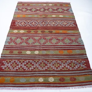 May include: Rectangular rug with a geometric pattern in shades of red, gray, green, and orange. The rug features horizontal stripes and diamond-shaped designs. The rug has a fringed edge.