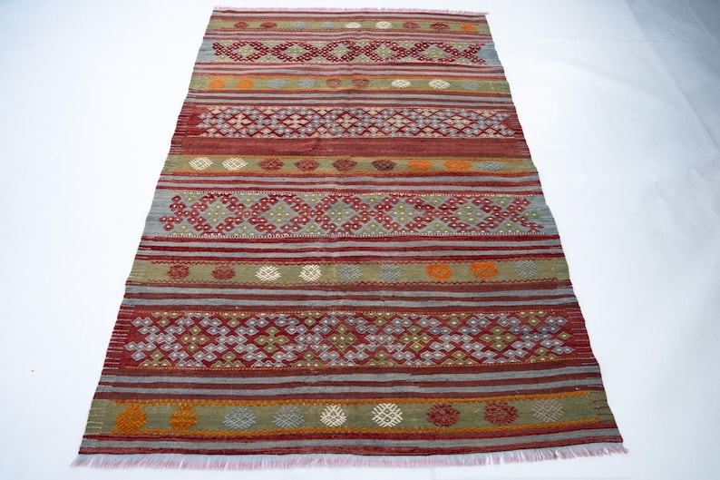 May include: Rectangular rug with a geometric pattern in shades of red, gray, green, and orange. The rug features horizontal stripes and diamond-shaped designs. The rug has a fringed edge.