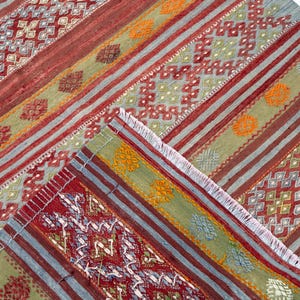 May include: A colorful, patterned rug with a geometric design. The rug features stripes in red, green, and blue, with intricate diamond and floral patterns. The rug has a fringed edge and is made of woven fabric.