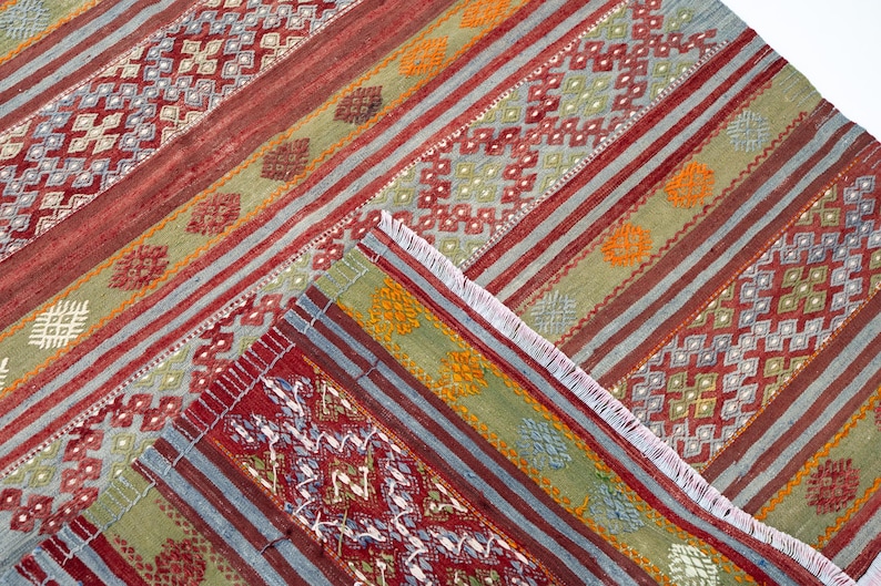 May include: A colorful, patterned rug with a geometric design. The rug features stripes in red, green, and blue, with intricate diamond and floral patterns. The rug has a fringed edge and is made of woven fabric.