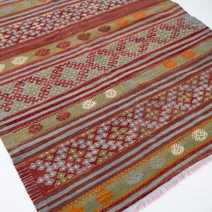 May include: A rectangular rug with a geometric pattern. The rug features alternating stripes of red, gray, and olive green. The design includes diamond and floral motifs, with orange and white accents. The rug has a fringed edge.