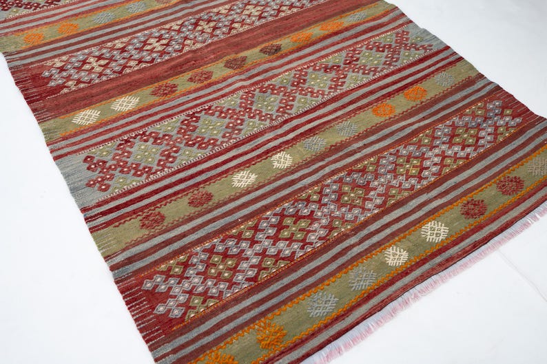 May include: A rectangular rug with a geometric pattern. The rug features alternating stripes of red, gray, and olive green. The design includes diamond and floral motifs, with orange and white accents. The rug has a fringed edge.