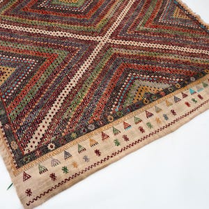 May include: A rectangular, hand-woven rug with a geometric pattern. The rug features a central diamond design with alternating stripes of red, green, and brown. The border is beige with colourful triangle and star motifs. Fringes are visible along the edges.