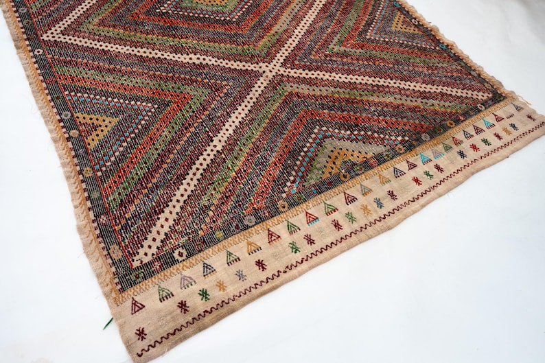 May include: A rectangular, hand-woven rug with a geometric pattern. The rug features a central diamond design with alternating stripes of red, green, and brown. The border is beige with colourful triangle and star motifs. Fringes are visible along the edges.