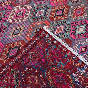 May include: A woven rug featuring a repeating geometric diamond pattern in vibrant shades of hot pink, teal, orange, and dark purple. A corner of the rug is folded over to reveal a deep red underside with intricate, multicolored woven details.