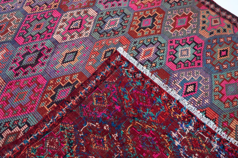 May include: A woven rug featuring a repeating geometric diamond pattern in vibrant shades of hot pink, teal, orange, and dark purple. A corner of the rug is folded over to reveal a deep red underside with intricate, multicolored woven details.
