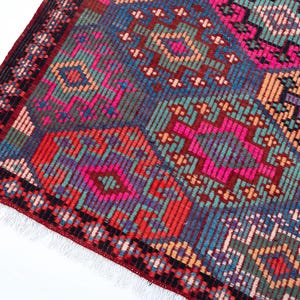 May include: A rectangular woven rug featuring a repeating geometric pattern of octagonal shapes in vibrant shades of hot pink, teal, deep red, orange, and purple. The rug has a dark red border with black accents and white fringe at the edge.