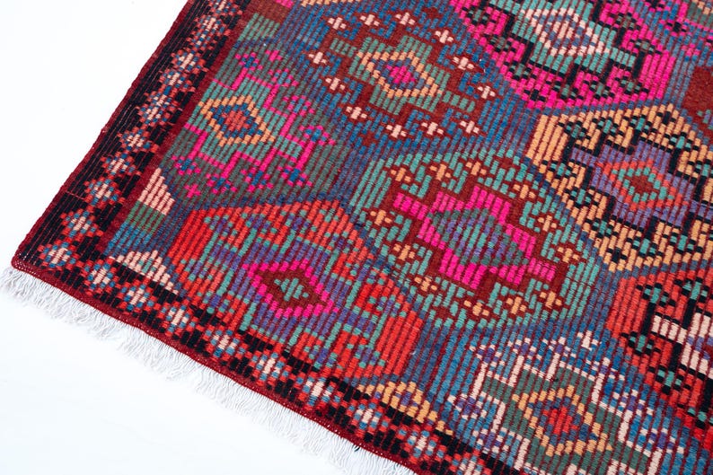 May include: A rectangular woven rug featuring a repeating geometric pattern of octagonal shapes in vibrant shades of hot pink, teal, deep red, orange, and purple. The rug has a dark red border with black accents and white fringe at the edge.