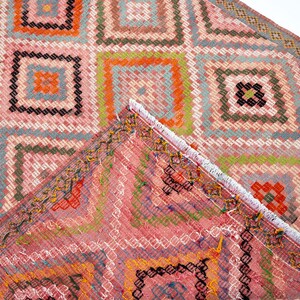May include: A close-up of a colorful, geometric patterned rug. The rug features a repeating square design in shades of pink, orange, green, and blue. The edges of the rug are visible, showing a fringed border.