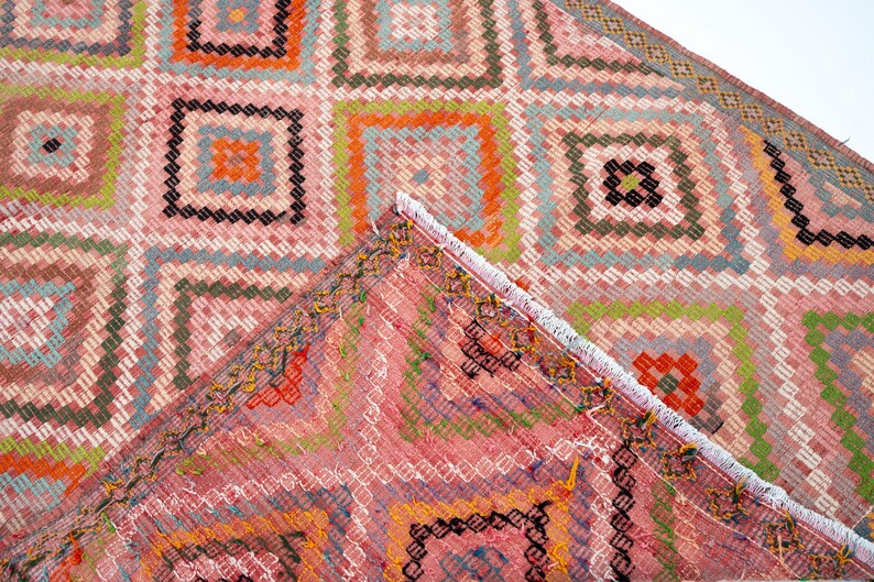 May include: A close-up of a colorful, geometric patterned rug. The rug features a repeating square design in shades of pink, orange, green, and blue. The edges of the rug are visible, showing a fringed border.