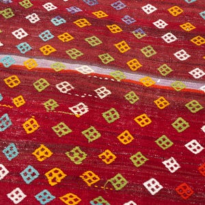 May include: Close-up of a red rug with a diamond pattern. The diamonds are in various colors, including white, yellow, green, and blue. The rug has a striped design with a light purple and orange stripe.