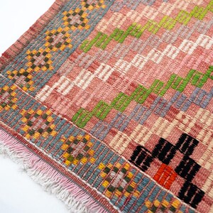 May include: Close-up of a woven rug with a pink and gray background. The rug features a geometric pattern with green, black, and orange accents. The edges are trimmed with a fringe.