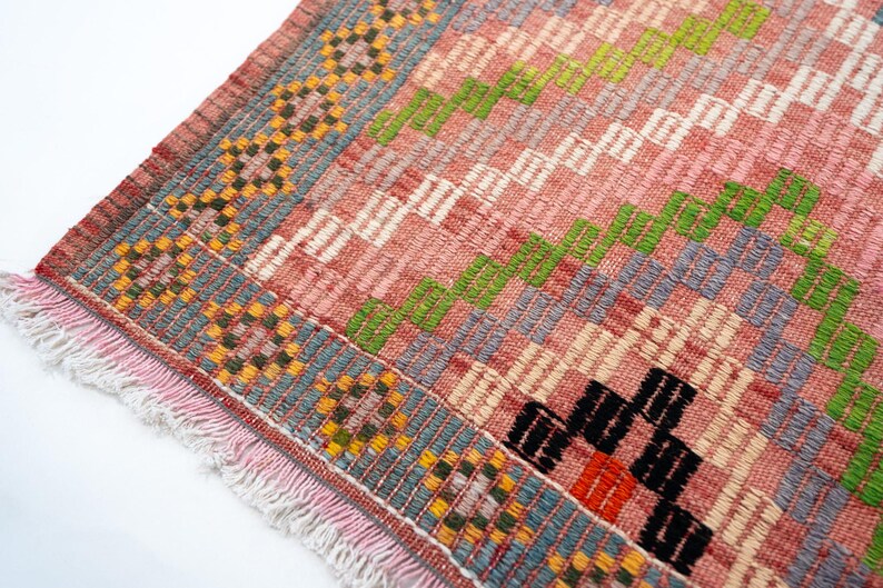 May include: Close-up of a woven rug with a pink and gray background. The rug features a geometric pattern with green, black, and orange accents. The edges are trimmed with a fringe.