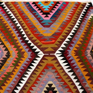 K&ouml;nnte beinhalten: Ein farbenfroher, geometrisch gemusterter Teppich mit einem zentralen Rautenmuster. Der Teppich weist eine Vielzahl von Farben auf, darunter Rot, Orange, Gelb, Gr&uuml;n, Lila und Wei&szlig;. Der Teppich besteht aus gewebtem Material und wird wahrscheinlich als Bodenbelag verwendet.