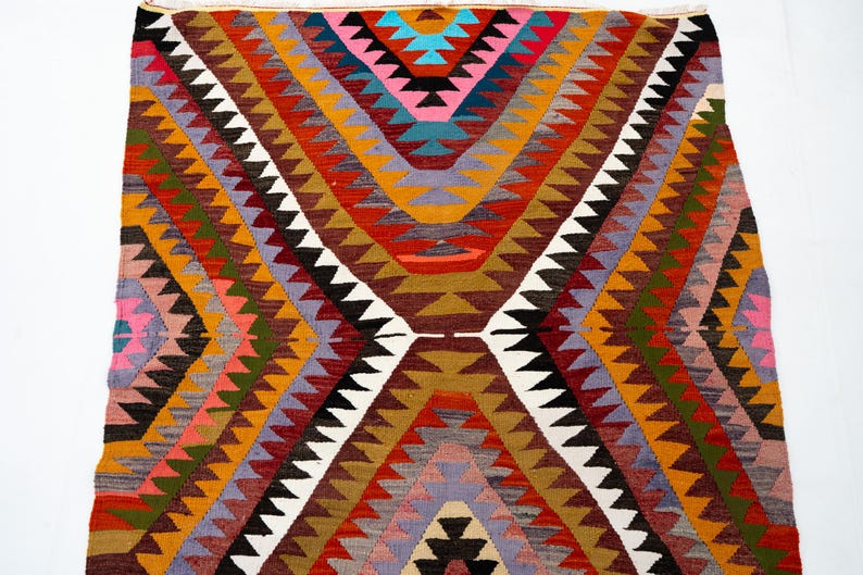 K&ouml;nnte beinhalten: Ein farbenfroher, geometrisch gemusterter Teppich mit einem zentralen Rautenmuster. Der Teppich weist eine Vielzahl von Farben auf, darunter Rot, Orange, Gelb, Gr&uuml;n, Lila und Wei&szlig;. Der Teppich besteht aus gewebtem Material und wird wahrscheinlich als Bodenbelag verwendet.