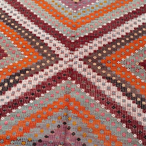May include: Close-up of a colorful woven rug with a geometric pattern. The rug features a repeating design of diagonal stripes in shades of orange, burgundy, gray, and white. Small dots of various colors are interspersed throughout the design.