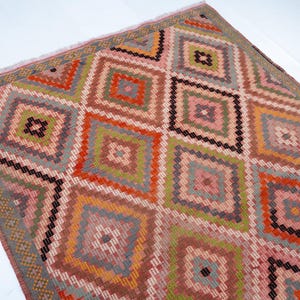 May include: A colorful, geometric rug with a diamond pattern. The rug features a variety of colors, including pink, orange, green, and gray. The rug has a fringed edge and is likely made of woven material.