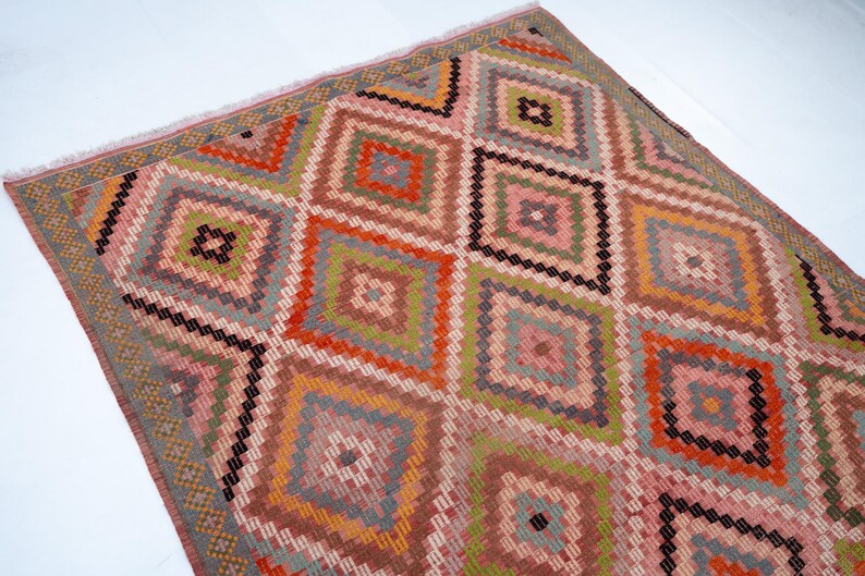 May include: A colorful, geometric rug with a diamond pattern. The rug features a variety of colors, including pink, orange, green, and gray. The rug has a fringed edge and is likely made of woven material.