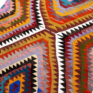 K&ouml;nnte beinhalten: Ein farbenfroher, geometrisch gemusterter Teppich mit einer Vielzahl von Farben, darunter Rot, Orange, Gelb, Blau und Lila. Der Teppich weist ein sich wiederholendes Rautenmuster mit wei&szlig;en Zickzack-Bord&uuml;ren auf. Der Teppich ist wahrscheinlich aus gewebtem Stoff.