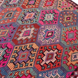 May include: A woven rug featuring a repeating geometric pattern of interconnected hexagons. The design incorporates a vibrant color palette of hot pink, teal, orange, purple, and deep red, with intricate stitched details forming diamond shapes.