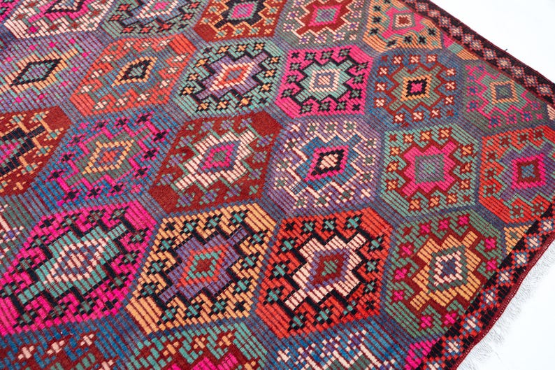 May include: A woven rug featuring a repeating geometric pattern of interconnected hexagons. The design incorporates a vibrant color palette of hot pink, teal, orange, purple, and deep red, with intricate stitched details forming diamond shapes.
