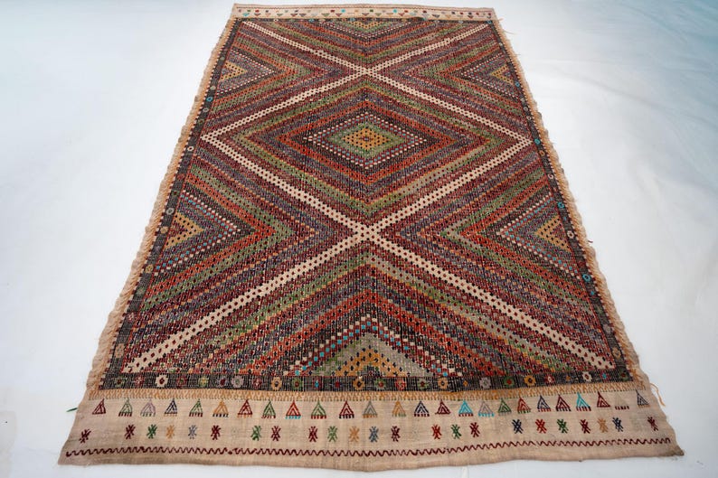 May include: Rectangular woven rug with a geometric diamond pattern in shades of red, green, brown, and cream. The rug has a cream-coloured border with small triangle and zigzag designs. The rug is approximately 152 cm long and 91 cm wide.