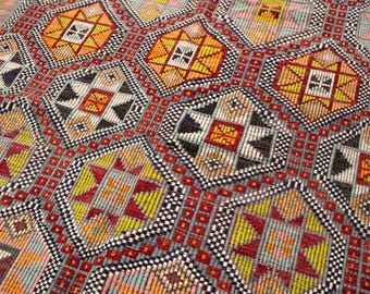 May include: Close-up of a colorful, patterned rug. The rug features geometric designs in shades of red, yellow, orange, black, and gray. The pattern includes diamond and square shapes, creating a textured appearance. The rug is likely made of woven fabric.