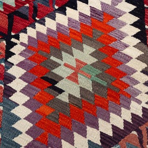 Puede incluir: Primer plano de un textil tejido con un patr&oacute;n geom&eacute;trico de diamantes. El dise&ntilde;o presenta una forma de diamante central en tonos de rojo, naranja, morado y blanco, rodeada por un borde rojo y azul. El textil es probablemente una alfombra o un tapiz.