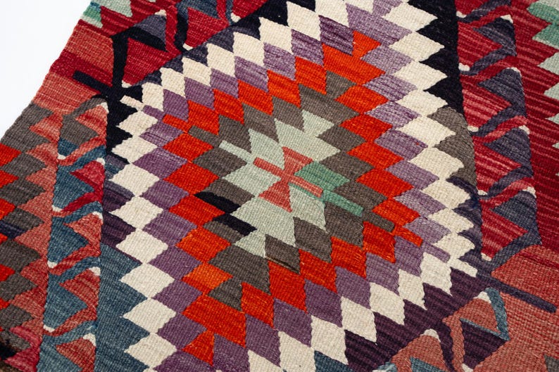 Puede incluir: Primer plano de un textil tejido con un patr&oacute;n geom&eacute;trico de diamantes. El dise&ntilde;o presenta una forma de diamante central en tonos de rojo, naranja, morado y blanco, rodeada por un borde rojo y azul. El textil es probablemente una alfombra o un tapiz.