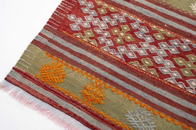 May include: Close-up of a woven rug with a geometric pattern. The rug features a red background with diamond-shaped designs in white, green, and gray. It has orange and gray stripes and a fringe edge. The rug is a traditional style.