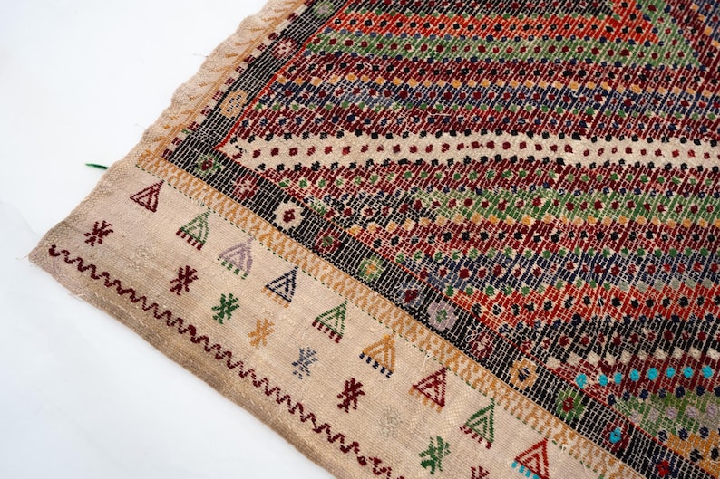May include: A section of a vibrant, patterned rug with a beige border. The rug displays a geometric design with red, green, blue, and black stripes. The border features triangle and star motifs in various colours. The rug has a fringed edge.