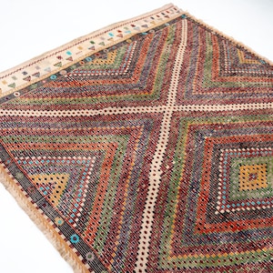 May include: A multi-coloured, geometric patterned rug with a diamond design. The rug has a cream border with small, colourful triangle and dot patterns. The main body of the rug features a repeating diamond pattern in shades of red, green, and orange.