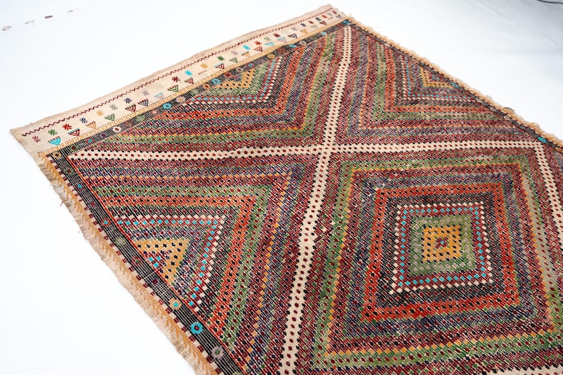 May include: A multi-coloured, geometric patterned rug with a diamond design. The rug has a cream border with small, colourful triangle and dot patterns. The main body of the rug features a repeating diamond pattern in shades of red, green, and orange.