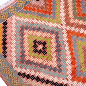 May include: Close-up of a colorful woven rug with a geometric pattern. The rug features a central diamond design in shades of pink, orange, green, and black, surrounded by a border of blue and yellow. The rug has a fringed edge.