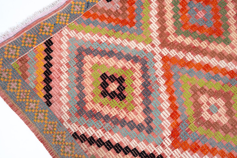 May include: Close-up of a colorful woven rug with a geometric pattern. The rug features a central diamond design in shades of pink, orange, green, and black, surrounded by a border of blue and yellow. The rug has a fringed edge.
